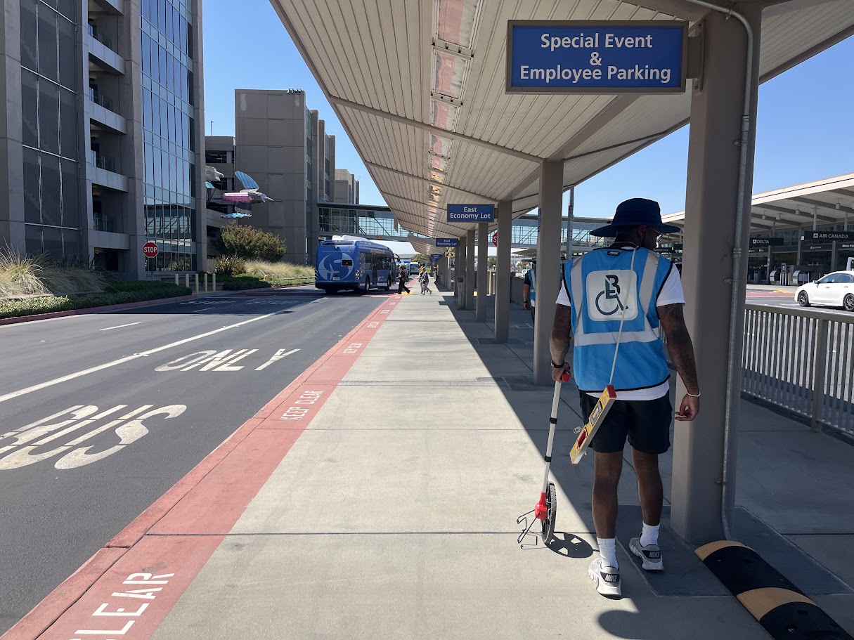 Accessibility inspector measuring a pedestrian route at an airport passenger loading area.