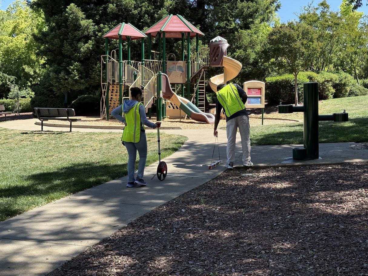 Two accessibility inspectors measuring a sidewalk route near a public playground.